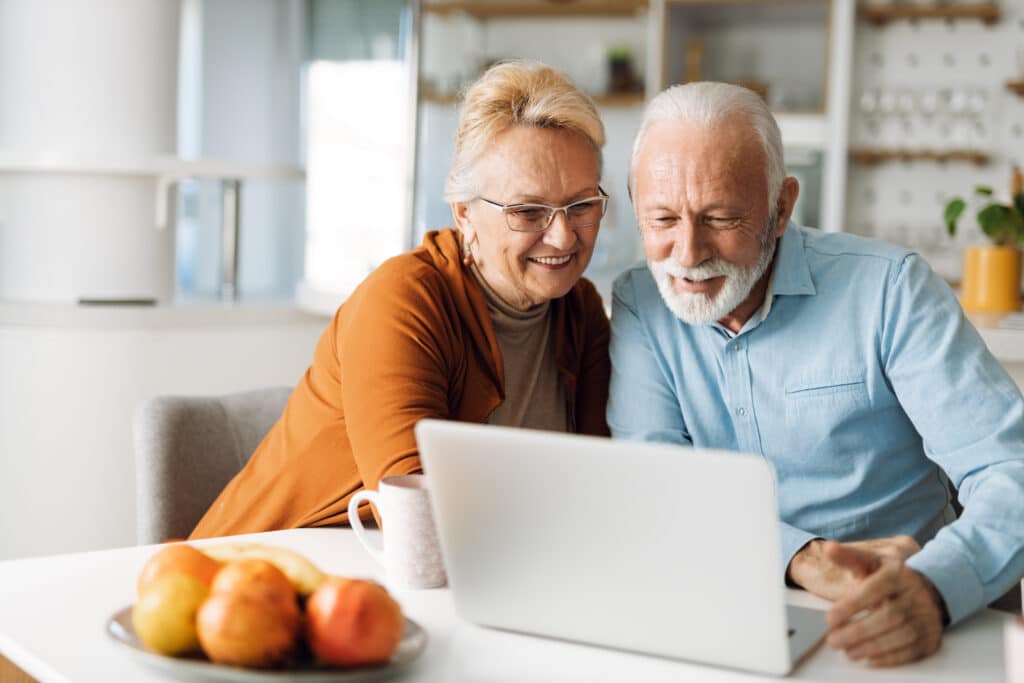 A joyful mature couple sitting together at home, smiling while using a laptop, exploring Hendersonville Assisted Living options.
