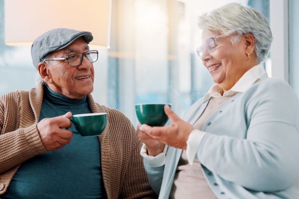Two seniors sit together on a couch, smiling and chatting over coffee in a bright, comfortable space, reflecting the everyday connection and comfort that come with senior routine benefits.