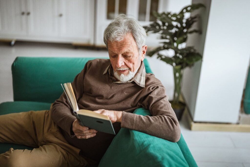 An older man settles into a comfortable sofa with a book, absorbed in a quiet afternoon that reflects the personal freedom and independence in senior living that lets residents spend their days exactly as they choose.