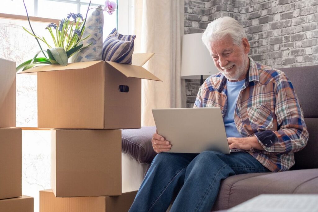 An older man sits smiling on a couch with his laptop, surrounded by stacked moving boxes topped with a plant and pillows — capturing the fresh-start feeling of retirement without homeownership, as he settles into a new, simplified chapter.